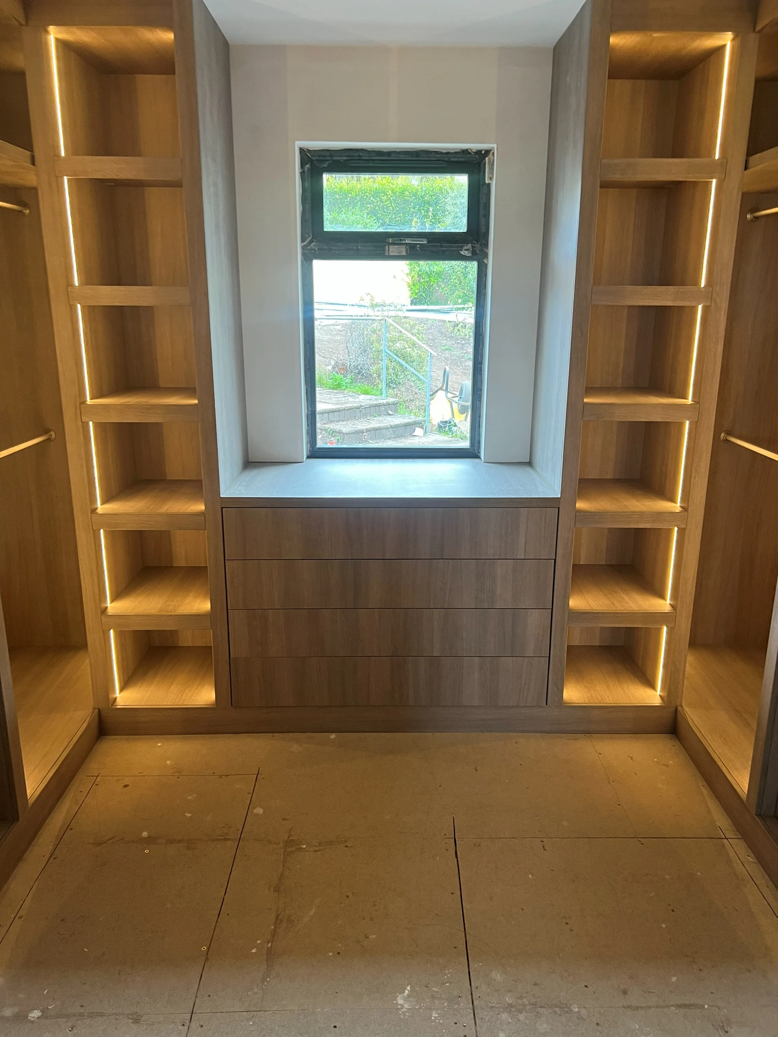 Installed oak dressing-room cabinetry centred on a window, with illuminated shelving and drawer storage